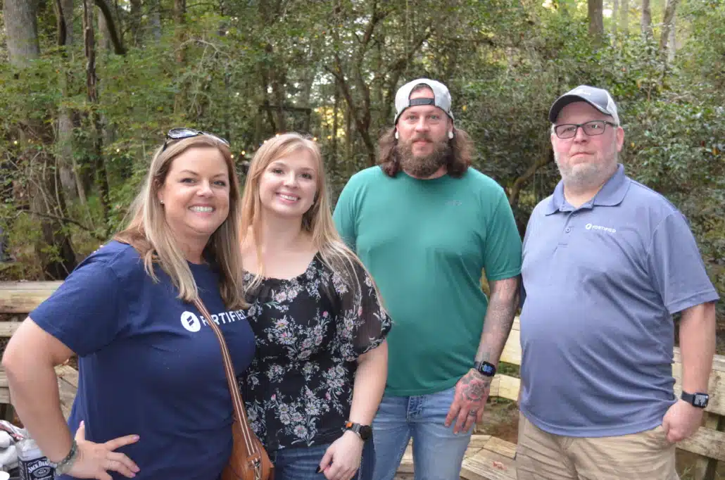 Four colleagues standing together on a wooden deck in the woods at a company retreat, two of them in Fortified Data shirts.