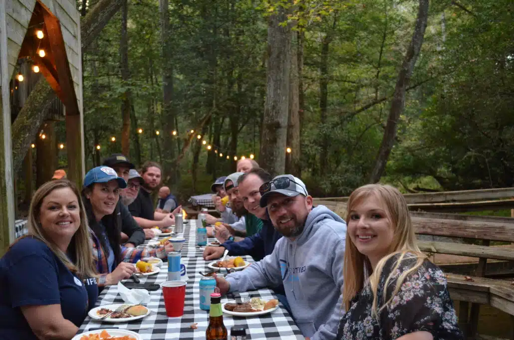 Colleagues eating at a long table under festoon lights at an outdoor pavilion in the woods.