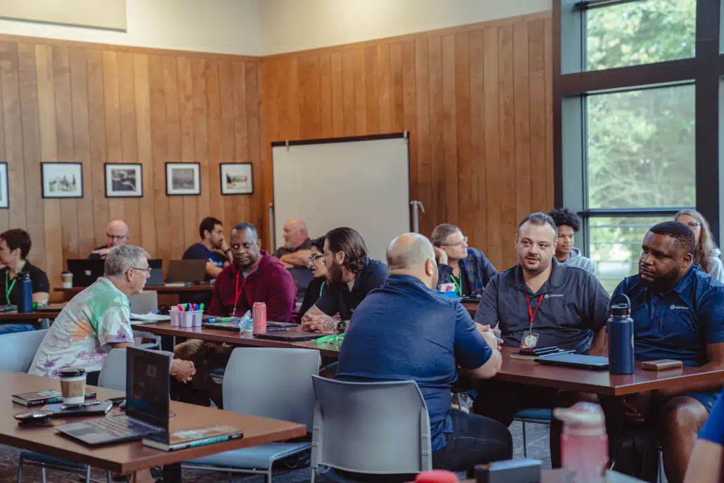 Colleagues working in small groups at tables in a wood-panelled meeting room, laptops and notebooks open.