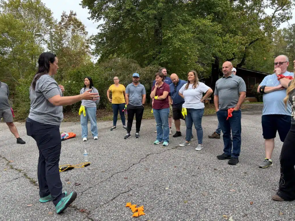 A group standing in a half circle on a paved clearing outdoors while one person explains a team exercise.