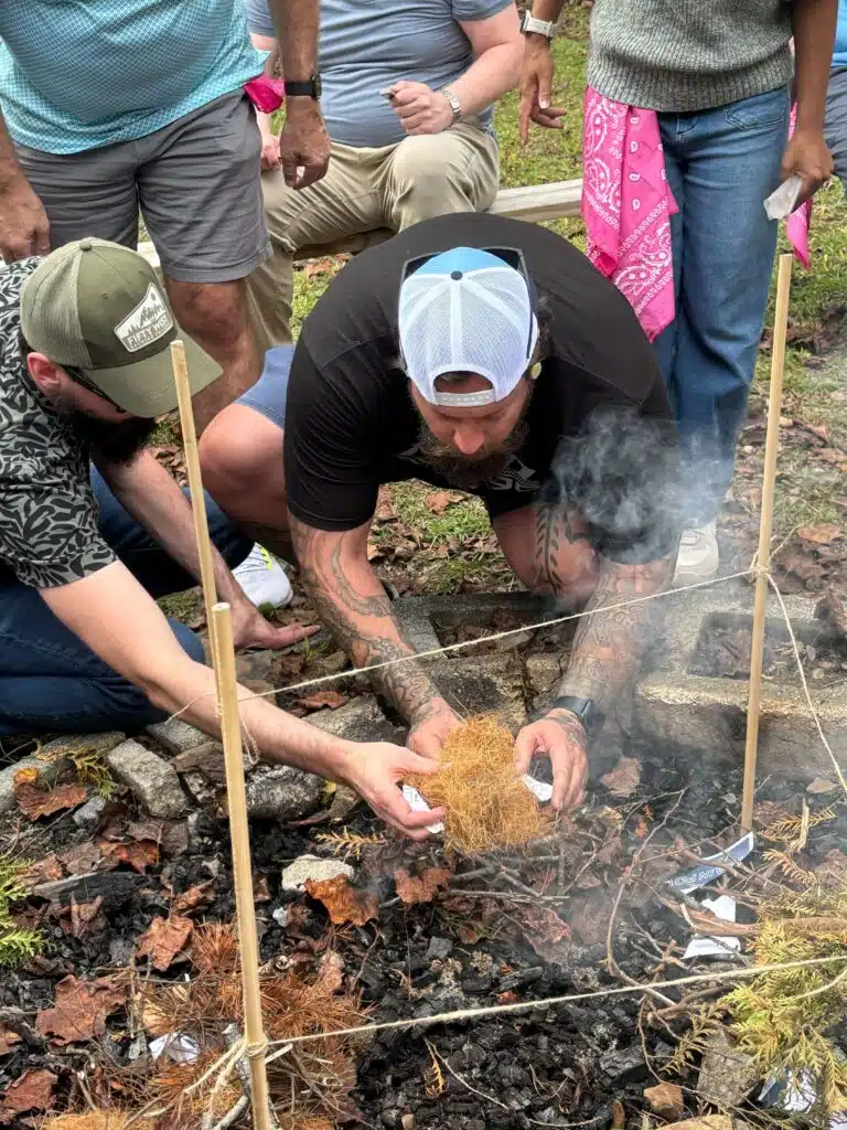 Two colleagues kneeling on the ground lighting a fire during an outdoor team challenge.