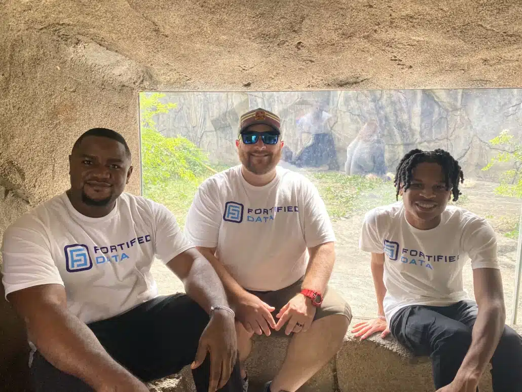Three colleagues in Fortified Data t-shirts sitting together in a shaded zoo viewing shelter.