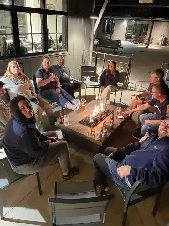 A dozen colleagues sitting in a circle around an outdoor gas fire pit in the evening.