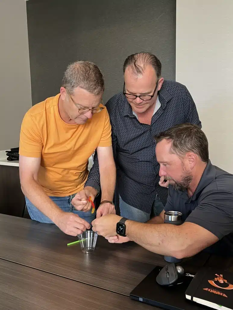 Three colleagues leaning over a table working on a hands-on exercise with a cup and a rubber band.