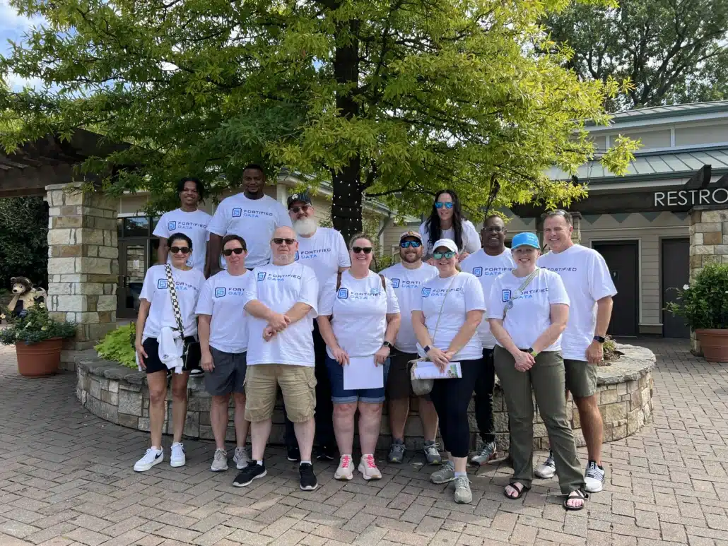 Fourteen colleagues in matching Fortified Data t-shirts posing outdoors on a paved courtyard under a tree.
