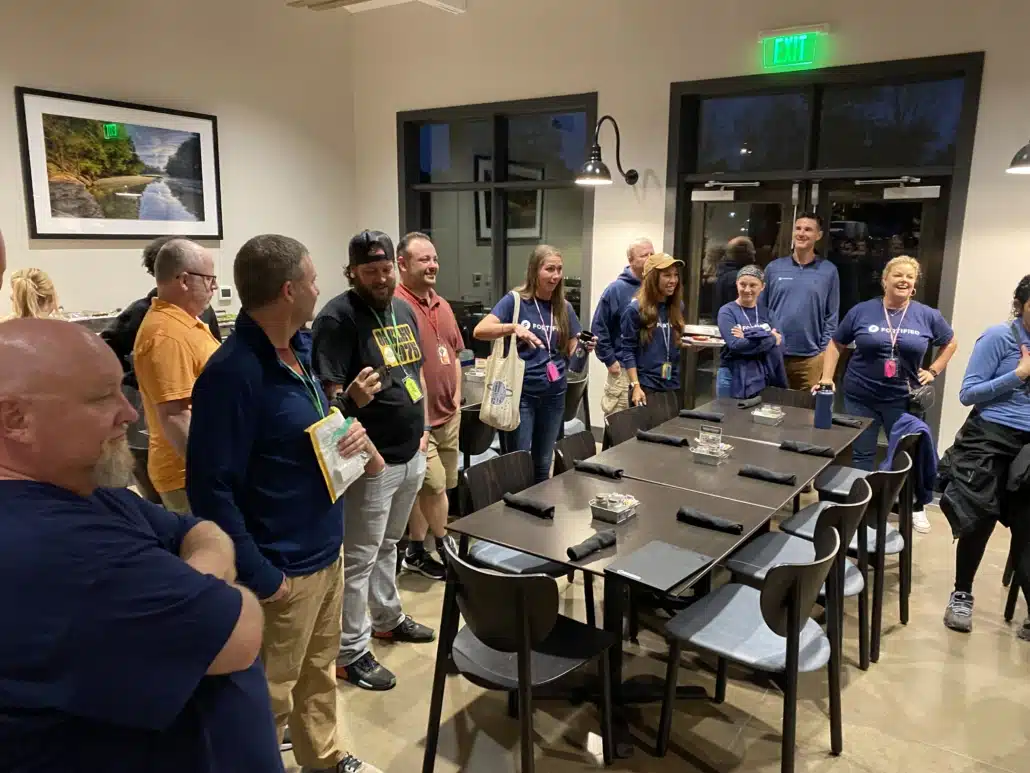 A group standing and talking beside a long laid dinner table in a lodge, several in Fortified Data shirts.