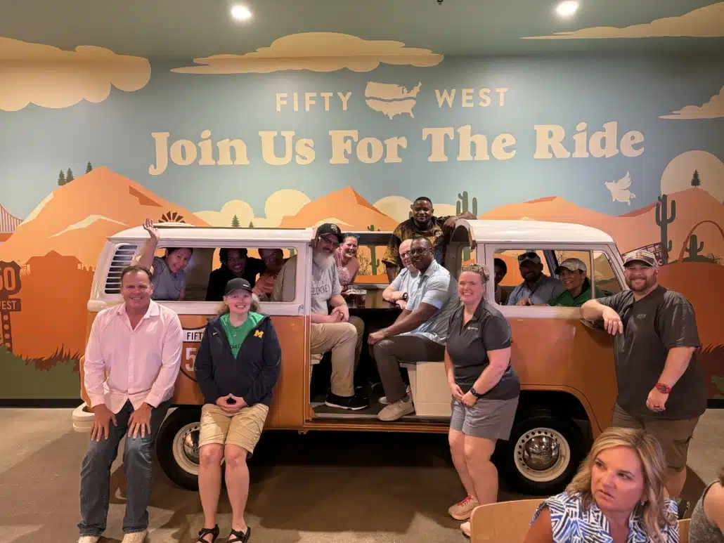 A large group posing in and around a vintage camper van in front of a painted mural reading Join Us For The Ride.